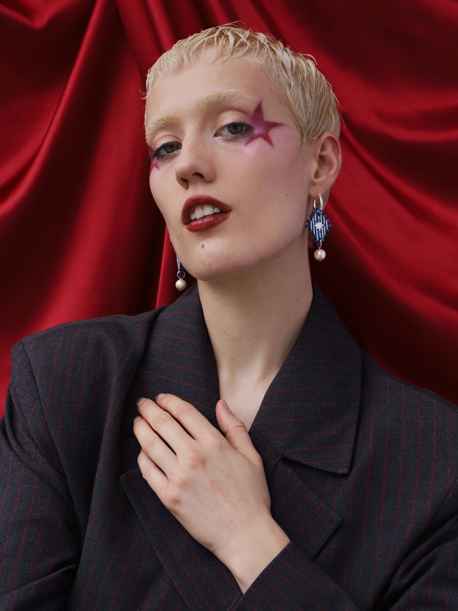 Model showcasing Cirque Hoops in Sapphire earrings with a striking makeup look against a red background.
