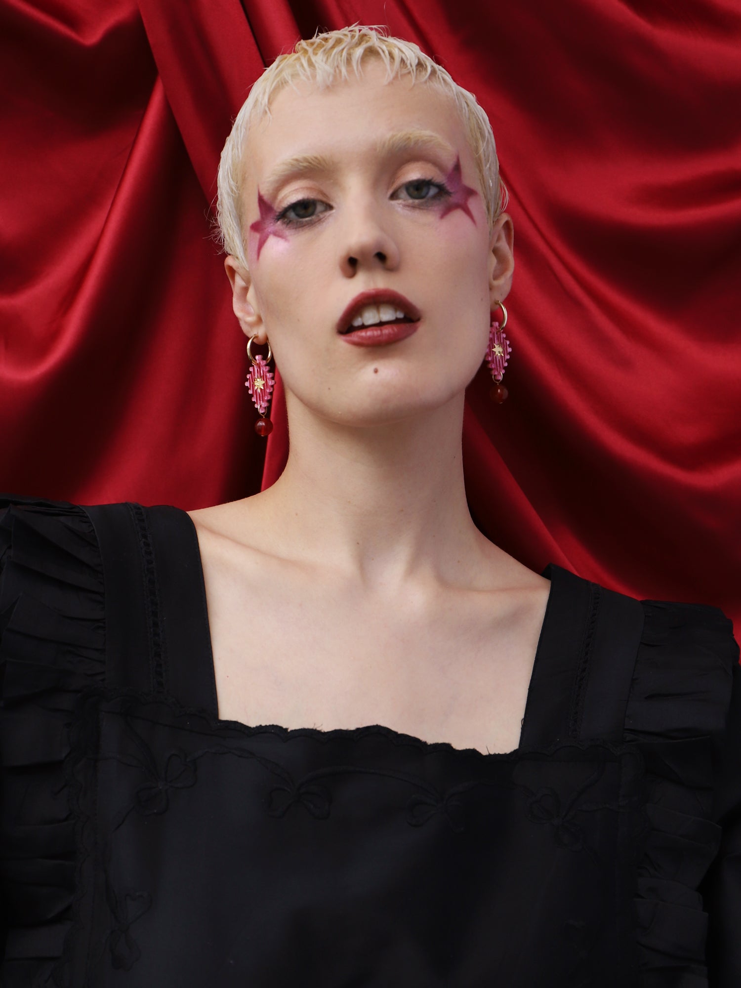 Model wearing Cirque Hoops in Rose earrings, showcasing unique design and vibrant style against a red backdrop.