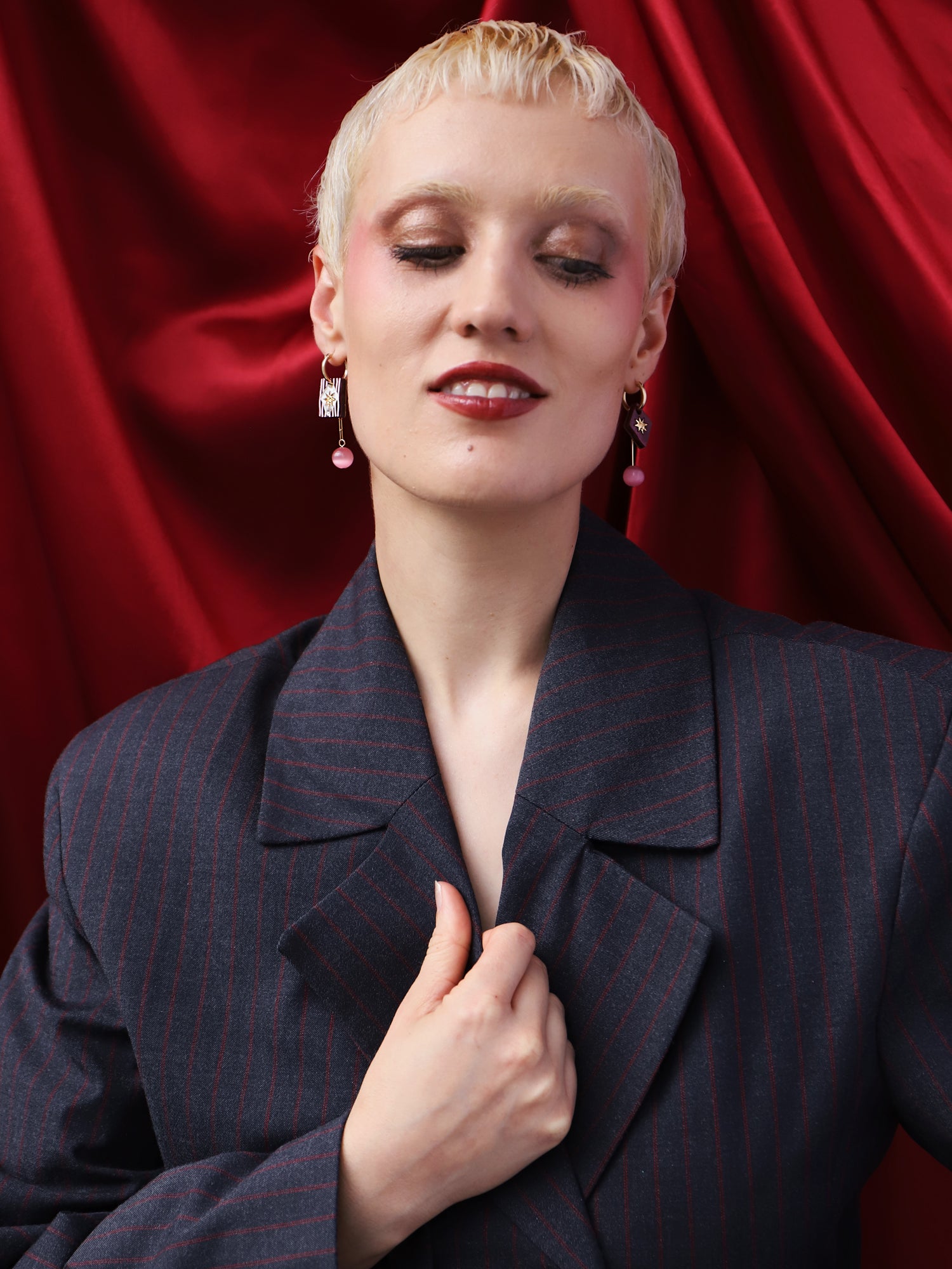 Model showcasing Stella Charm Hoops in Cherry earrings, styled with a chic oversized blazer against a red backdrop.