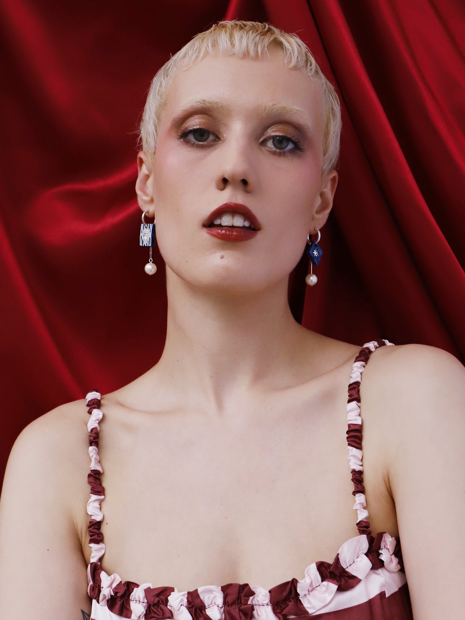 Model wearing Stella Charm Hoops in Sapphire earrings against a red backdrop, showcasing handmade design and charm details.